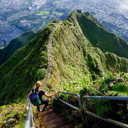 A female hiker on vacation climbing the Haiku Stairs Stairway to Heaven over a thin ledge on the green mountain in Oahu, Hawaii which was made possible by Sky Bird Travel & Tours customized vacation packages.